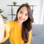 Pretty young asian female with big smile sitting at living room. She having fun taking light cheerful selfie on blurred background