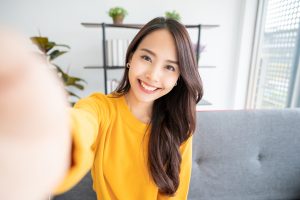 Pretty young asian female with big smile sitting at living room. She having fun taking light cheerful selfie on blurred background