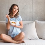 Happy young woman eating healthy breakfast while sitting on a couch at home