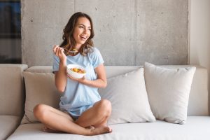 Happy young woman eating healthy breakfast while sitting on a couch at home