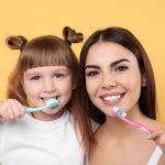 Little girl and her mother brushing teeth together on color background