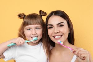 Little girl and her mother brushing teeth together on color background