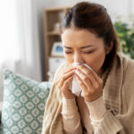 healthcare, cold and people concept - sick asian woman in blanket taking paper tissue from box and blowing her nose at home